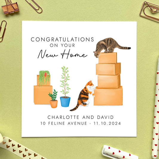 A personalised congratulations on your new home card. The design features a brown tabby cat on top of a pile of boxes, looking down at a calico cat. It can be customised with names, address and moving date.