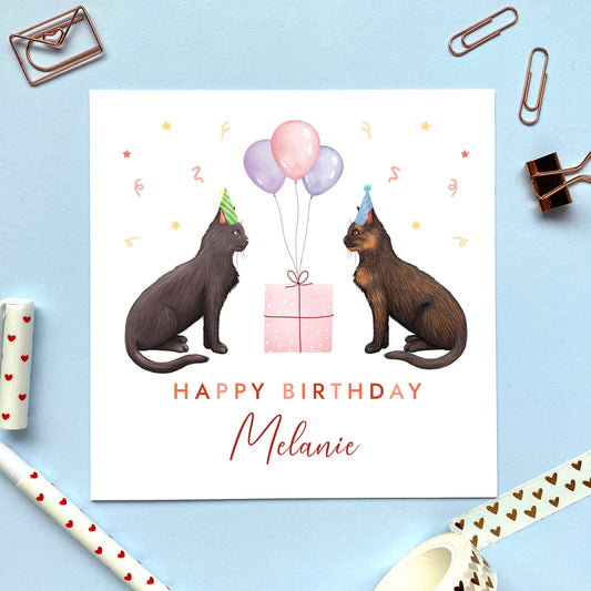 A personalised two cats birthday card for her where you can choose both cats on the card and add an age and name. The card in the photo is a birthday card with a black cat and a tortoiseshell (black and ginger) cat.