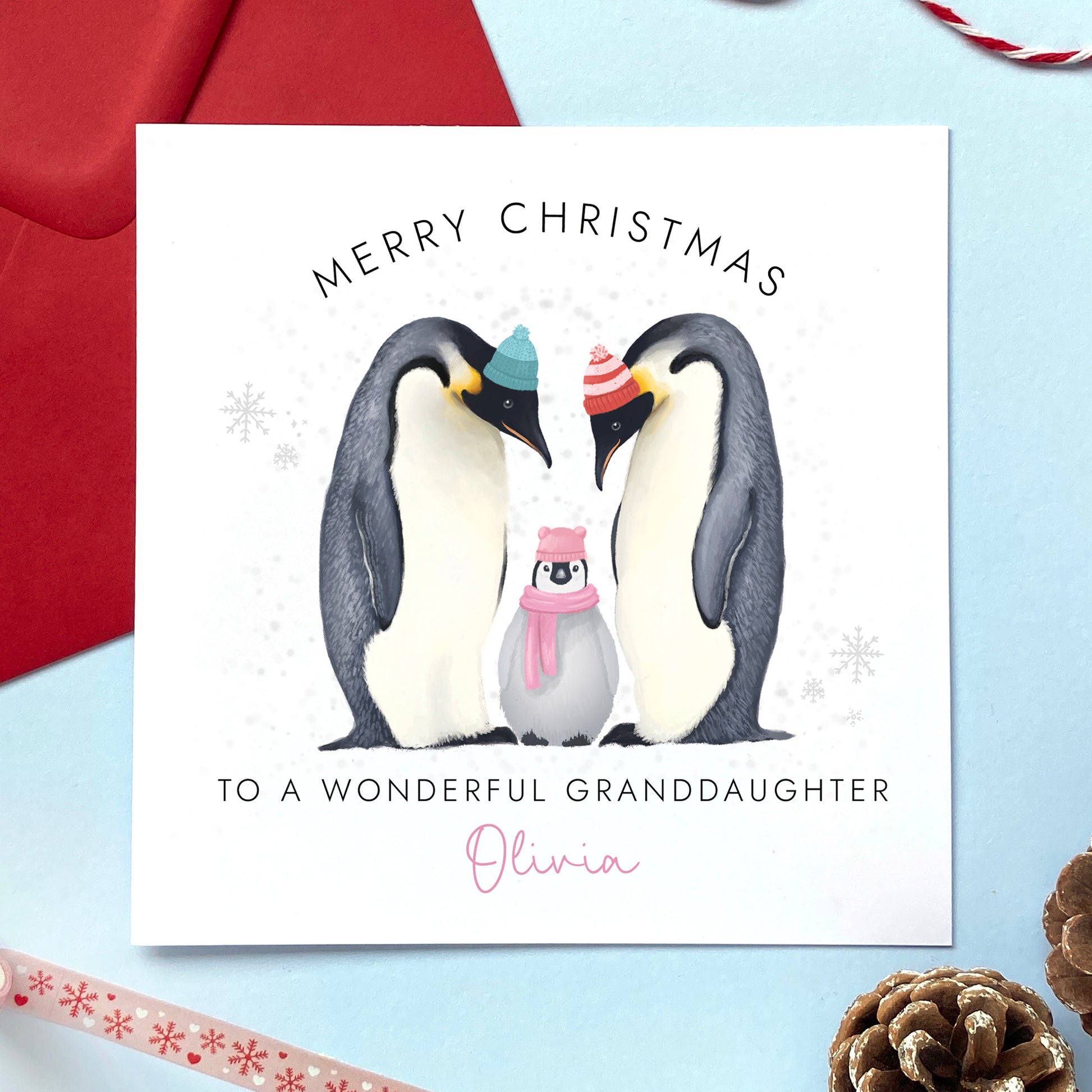 A personalised Christmas Card for Girl featuring two adult penguins and a baby penguin wearing a pink scarf. It can be personalised with a name. The card in the photo is a Christmas Card for Granddaughter.