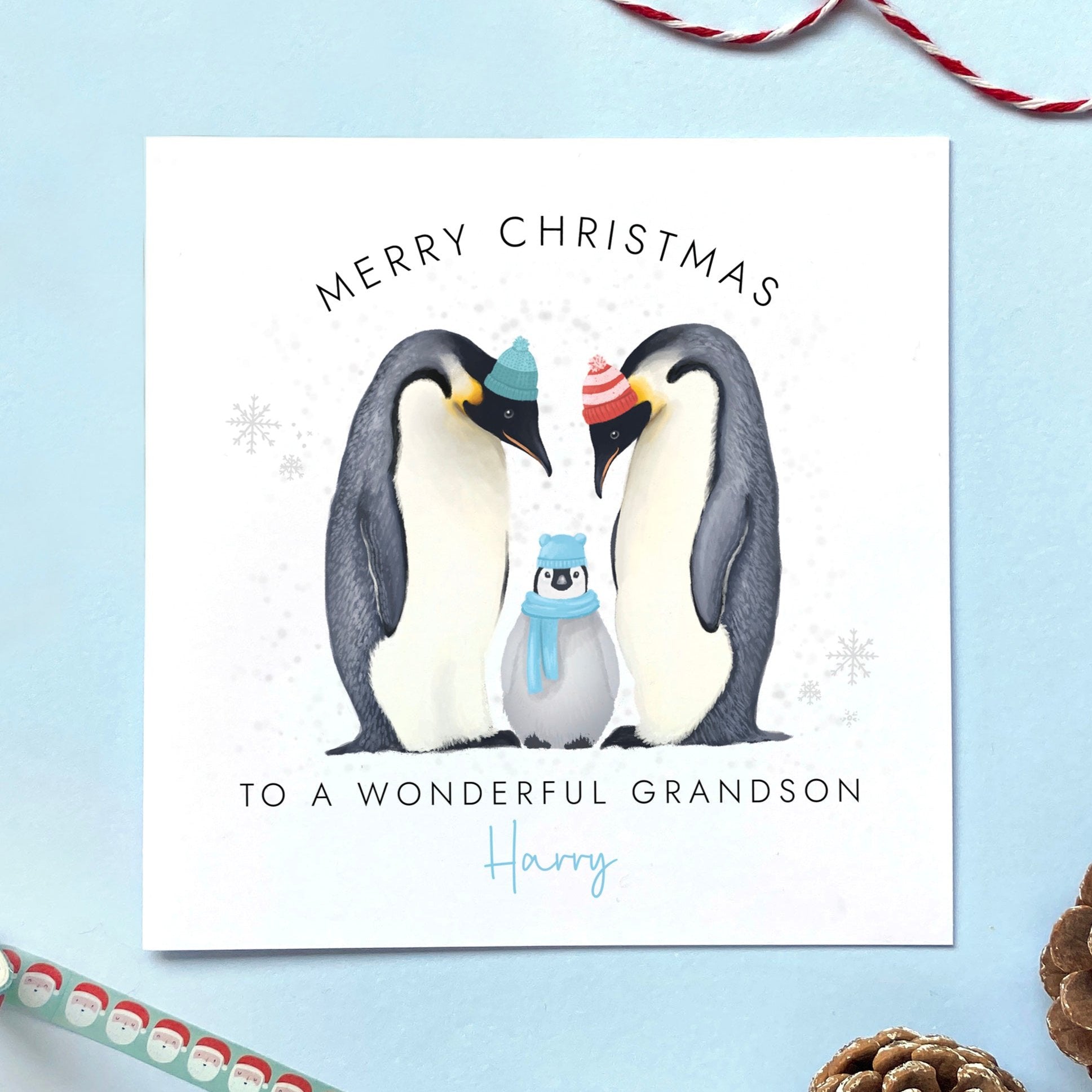 A personalised Christmas Card for Grandson featuring two parent penguins and a baby penguin wearing a blue scarf. It can be customised with a name. The card in the photo is a Christmas Card for Grandson.