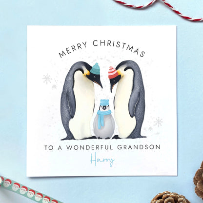 A personalised Christmas Card for Grandson featuring two parent penguins and a baby penguin wearing a blue scarf. It can be customised with a name. The card in the photo is a Christmas Card for Grandson.