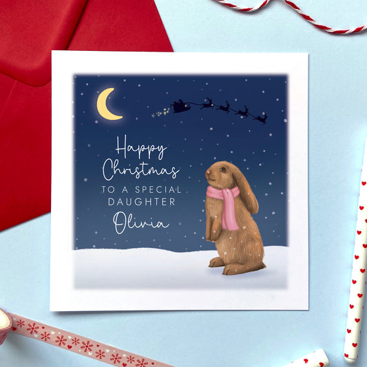 A personalised Christmas card for Daughter featuring a brown bunny rabbit sitting in the snow, wearing a pink scarf. The card in the photo is a Christmas Card for Daughter.