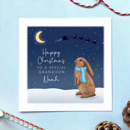 A personalised Christmas card for Boy featuring a brown bunny rabbit sitting in the snow, wearing a blue scarf. The card in the photo is a Christmas Card for Son.