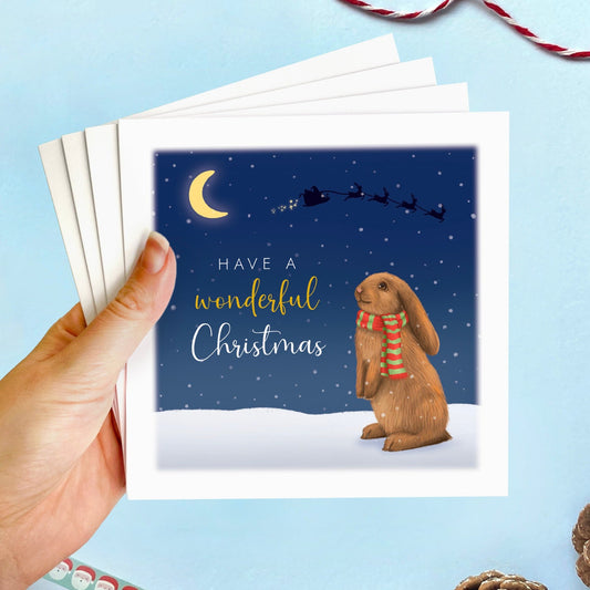 A Christmas Card featuring a Brown Bunny Rabbit wearing a scarf. The background is a snowy nighttime scene with a silhouette of Santa in the sky. It comes in packs of 4 cards, 8 cards and 12 cards.